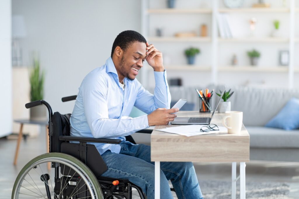 Remote job for disabled people. Happy black guy in wheelchair communicating online, using smartphone